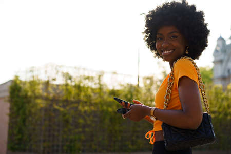 Smiling afro-american woman holding mobile phone while walking in the street