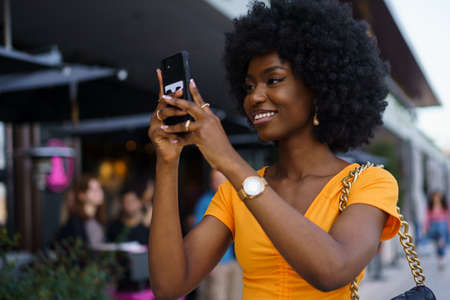 Smiling afro-american woman holding mobile phone while walking in the streetの写真素材