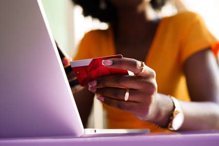 Happy young african woman holding credit card while shopping via laptop sitting in outdoor cafe