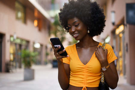 Smiling afro-american woman holding mobile phone while walking in the streetの写真素材