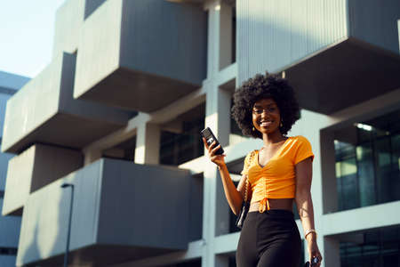 Smiling afro-american woman holding mobile phone while walking in the street
