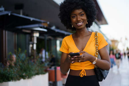Smiling afro-american woman holding mobile phone while walking in the street