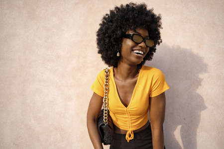 Happy African young woman with afro hair wearing stylish sunglasses posing against beige wall