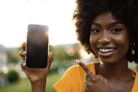 Portrait of a smiling young african woman holding blank screen mobile phone standing in the street