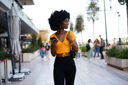 Smiling afro-american woman holding mobile phone while walking in the streetの写真素材