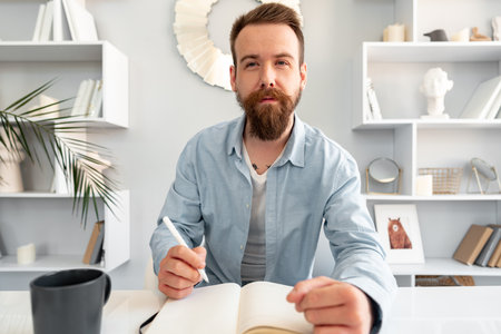 Young bearded man sitting at the desk and taking notes at homeの写真素材
