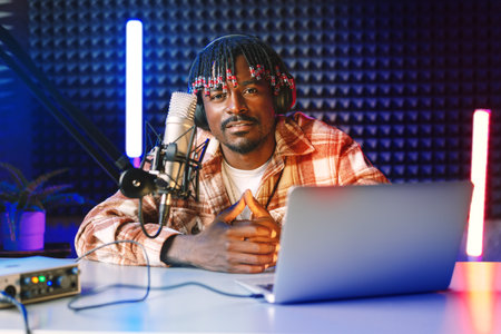 African radio host sitting at desk recording in studio with microphone and laptopの写真素材
