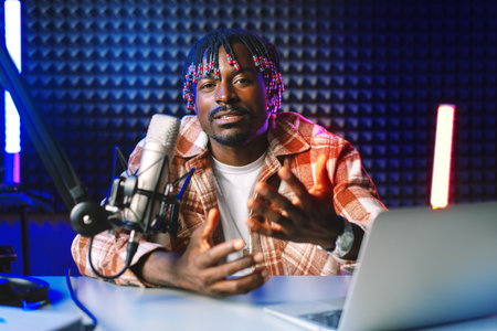 African radio host sitting at desk recording in studio with microphone and laptopの写真素材
