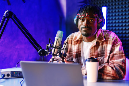 African radio host sitting at desk recording in studio with microphone and laptopの写真素材
