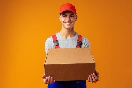 Young courier in uniform with box on yellow background in studioの写真素材