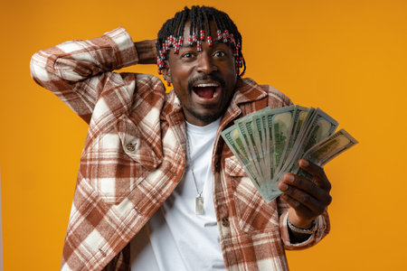 Young happy rich african man in casual shirt holding dollar bills against yellow backgroundの写真素材