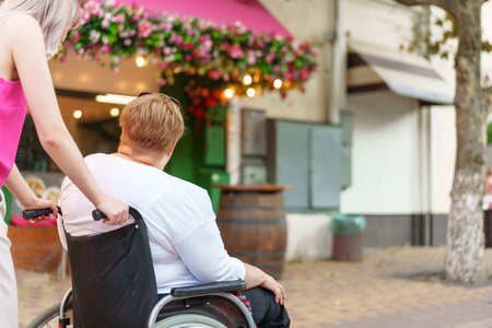 Back view of young woman helping mature woman in wheelchair in the cityの写真素材
