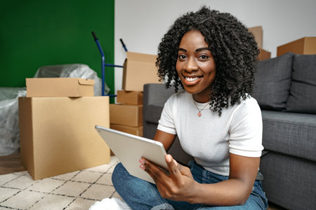 Young African woman using a tablet as she sits in living room surrounded by cardboard boxes
