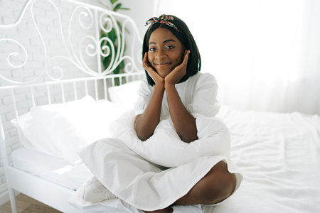 Beautiful young thoughtful african woman sitting in morning bedの写真素材