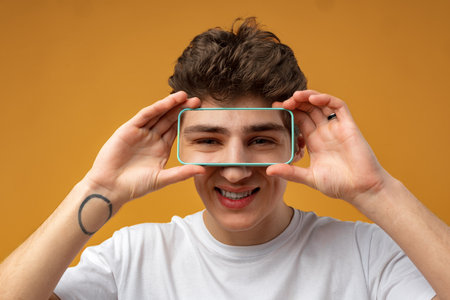 Young man covering his eye with mobile phone over yellow background.