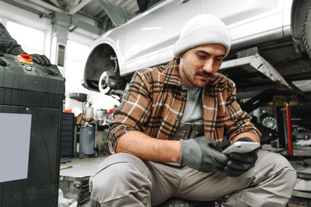 Car service worker sitting on a wheel and resting, using mobile phoneの写真素材