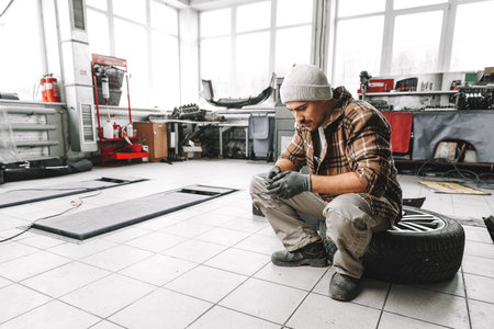 Car service worker sitting on a wheel and resting, using mobile phoneの写真素材