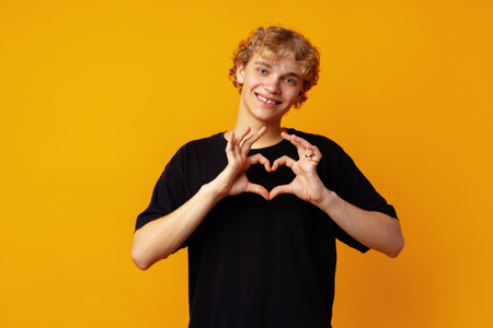 Young man on yellow background smiling and showing a heart shape with handsの写真素材