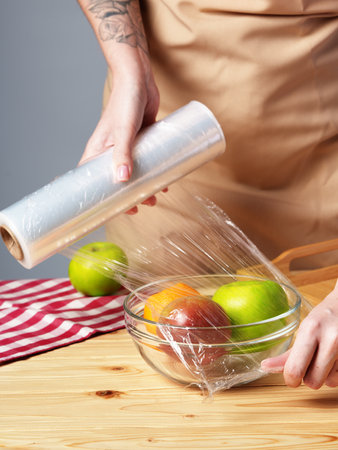 Woman putting cling filmover bowl of fresh fruit at wooden tableの写真素材