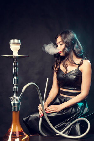Young beautiful female in black dress smoking and exhales hookah