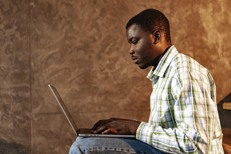 Young african man sitting on a wooden ladder with laptop in a room