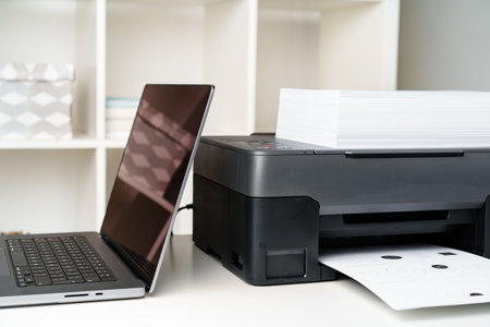 Modern printer with paper and laptop on table in office