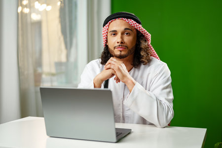 Young Arab man in traditional dress thobe working on computer in officeの写真素材