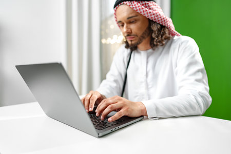 Young Arab man in traditional dress thobe working on computer in officeの写真素材