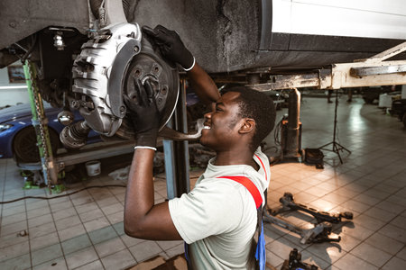 African male auto-mechanic repairing car brakes under the car in auto serviceの写真素材
