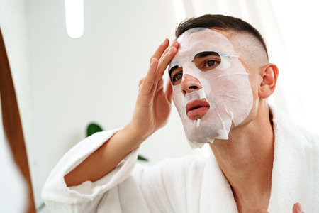Handsome young man applying sheet mask near mirror in bathroomの写真素材