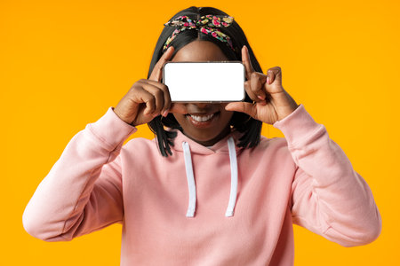 Portrait of a excited young african woman showing blank screen mobile phone over yellow backgroundの写真素材