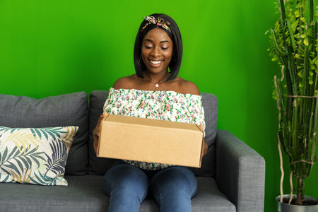 African young woman sit on couch at home unpack cardboard box with online purchaseの写真素材