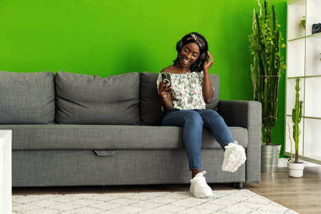 African woman listening to music with headphones while sitting on couch at homeの写真素材