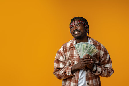 Young happy rich african man in casual shirt holding dollar bills against yellow background