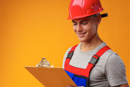 Young construction worker wearing hardhat and holding a clipboard on yellow background in studioの写真素材