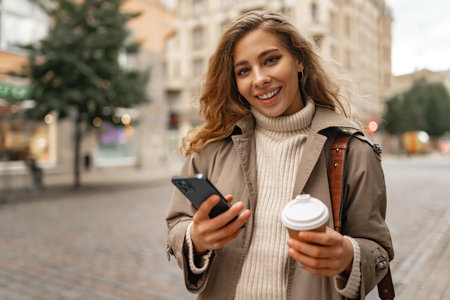 Young woman with curly blonde hair using the phone with a cup of coffee in hands on the city streetsの写真素材