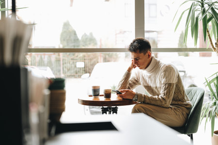 Young Man Engrossed in Smartphone at a Modern Cafe During Daytimeの写真素材