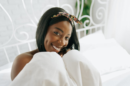 Beautiful young thoughtful african woman sitting in morning bedの写真素材