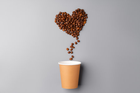 Coffee beans arranged in a heart shape above a paper cup on a grey background