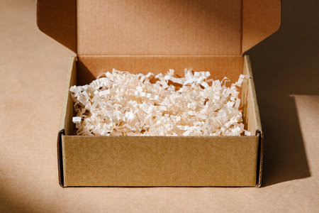 Open cardboard box with shredded paper packaging resting on a light brown surface illuminated by soft natural lightの写真素材