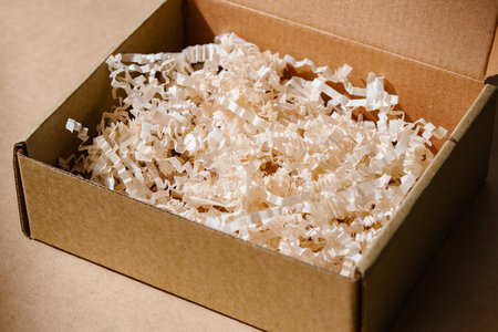 Open cardboard box with shredded paper packaging resting on a light brown surface illuminated by soft natural lightの写真素材