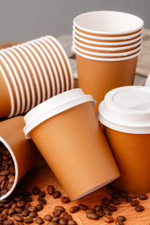 Brown disposable coffee cups arranged with coffee beans and a newspaper on a wooden surface in a casual settingの写真素材