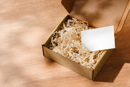 Open cardboard box filled with shredded paper and a blank card on a wooden surfaceの写真素材