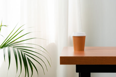 A disposable coffee cup on a wooden table beside a leafy plantの写真素材