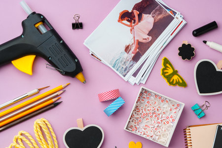 Crafting supplies arranged on a pink background, including glue gun, scissors, and decorative items...