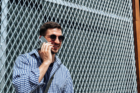 A man in a blue checkered shirt smiles while standing outside a modern building on a sunny day