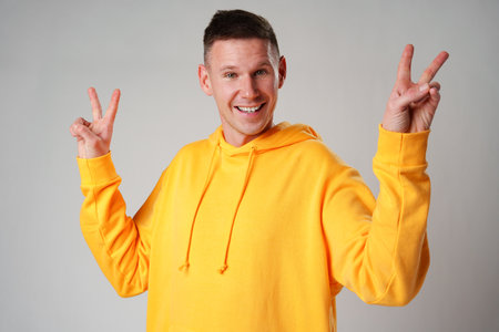 Smiling young man in yellow hoodie showing victory sign against gray background