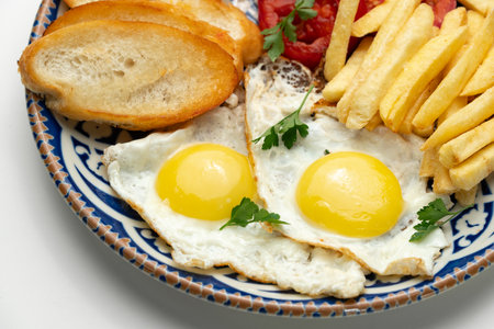 Breakfast plate with fried eggs, sausage, tomatoes, and fries served on a decorative blue plate