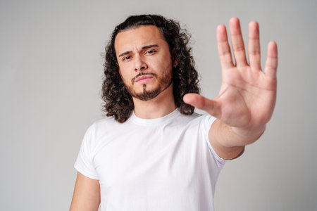 Young man with curly hair gestures to stop while wearing a plain white t-shirt against a neutral backgroundの写真素材