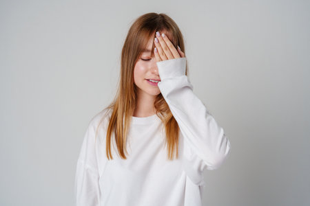 A young woman in a white oversized sweater poses thoughtfully against a plain backdrop while partially covering her faceの写真素材